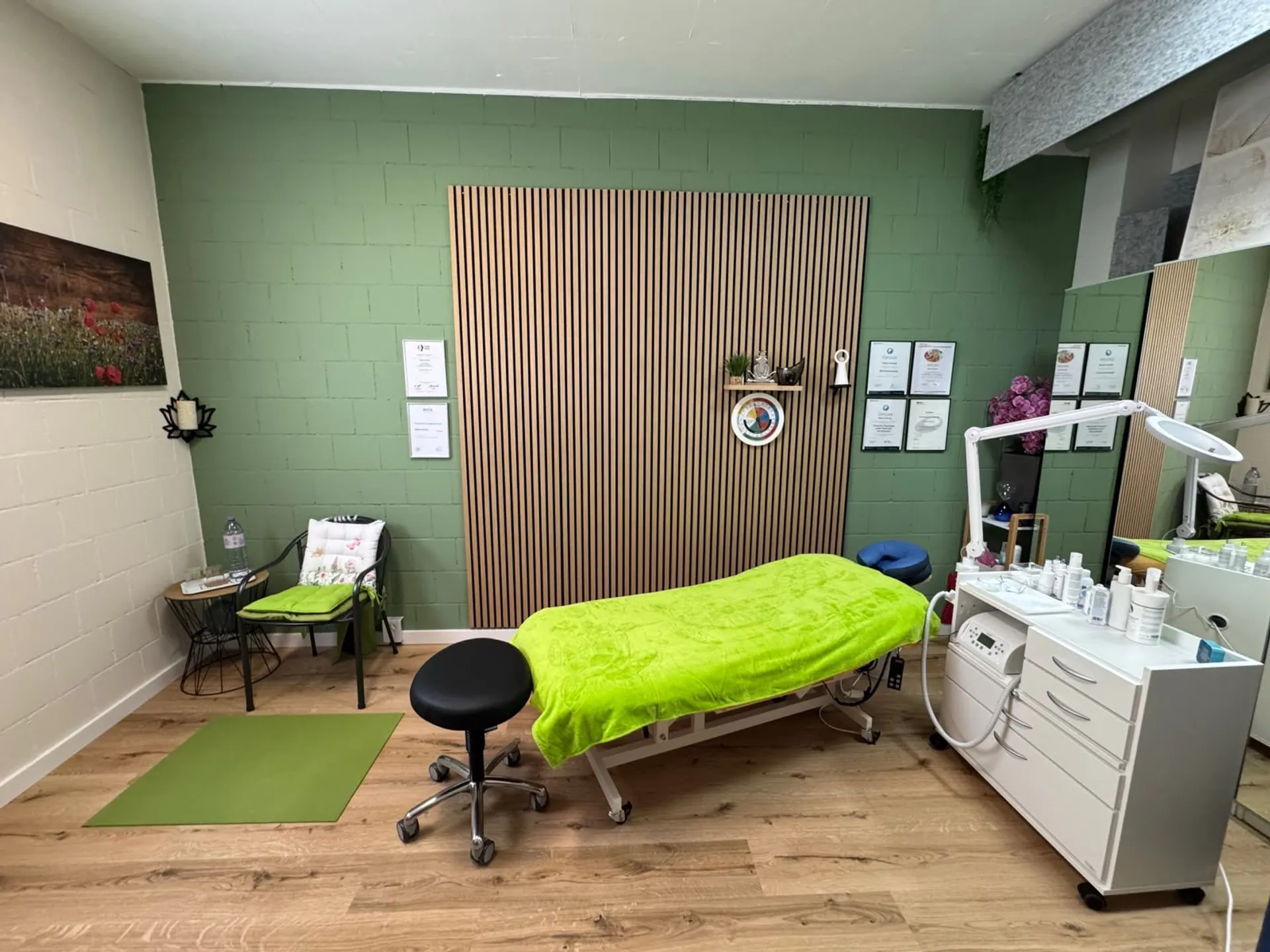 Behandlungsraum der Praxis: Massageliege mit grünem Tuch vor salbeigrüner Wand und Holzlamellen-Paneel, daneben Stuhl und Yogamatte